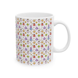 Könnte beinhalten: Eine weiße Keramiktasse mit einem bunten Muster aus Comicfiguren. Die Figuren sind in verschiedenen Farben, darunter Rot, Orange, Gelb, Grün, Blau, Lila und Pink. Die Figuren lächeln alle und haben kleine, runde Körper. Die Tasse hat einen weißen Henkel.
