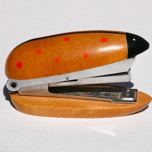 May include: A wooden stapler featuring a smooth, light brown finish with a black painted tip and small red circular accents. The stapler has a metallic silver mechanism integrated between the curved wooden top and the solid wooden base.
