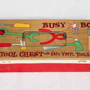 May include: A rectangular metal tool chest with a tan lid and red base. The lid features the text BUSY BOY TOOL CHEST with SAFE VINYL TOOLS, a cartoon illustration of a child, and colorful graphics of various tools including a hammer and saw.