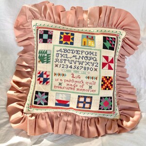 Könnte beinhalten: Ein quadratisches Zierkissen mit einem altrosa Rüschenrand. Die Mitte besteht aus cremefarbenem Stoff mit einem Kreuzstich-Mustertuch-Design, das ein Alphabet, Zahlen, kleine Quiltblock-Motive und den Text enthält: Life is a patchwork quilt made of treasured memories. Linda 1989 Lee ist unter den Zahlen eingestickt.