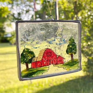Op de afbeelding: Een rechthoekige glas-in-lood zonnevanger met een handgeschilderde scène van een felrode schuur in een groen landschap met twee bomen. Het stuk is omlijst met een donkere metalen rand en hangt aan een dun touwtje.