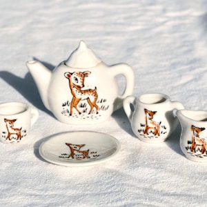 May include: A miniature ceramic tea set featuring a white glaze with hand-painted brown and tan deer illustrations. The set includes a teapot with a lid, a small cup, a saucer, a sugar bowl with two handles, and a creamer pitcher with a handle.