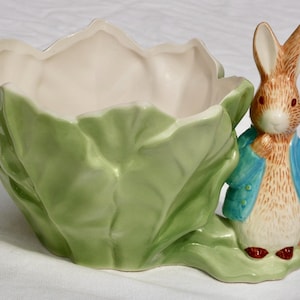 May include: A ceramic bowl shaped like a green cabbage leaf with a sculpted figure of a brown rabbit wearing a blue jacket attached to the side. The rabbit has long ears, dark eyes, and is holding a small orange carrot against its side.
