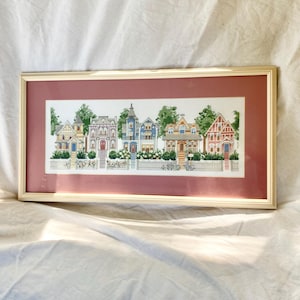 May include: A framed cross-stitch embroidery depicting a row of five colorful Victorian-style houses with detailed architecture, green trees, and a white picket fence, set against a deep rose-colored matting within a light-colored wooden frame.