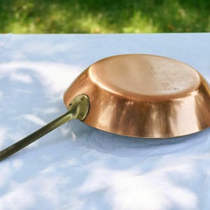 May include: A round copper frying pan with a polished metallic finish and a long, slender brass handle attached by a riveted plate. The pan is positioned upside down, showcasing its smooth, curved exterior and the sturdy metal construction.