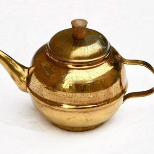 May include: A polished brass teapot featuring a rounded body, a curved spout, and a handle. The lid is topped with a small wooden knob. The surface of the teapot is decorated with etched leaf patterns and fine cross-hatched textures.