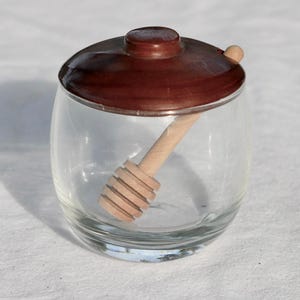 May include: A clear glass honey pot with a rounded body and a smooth, dark brown wooden lid. A light-colored wooden honey dipper with a multi-grooved head is positioned inside the jar, resting at an angle against the interior glass wall.