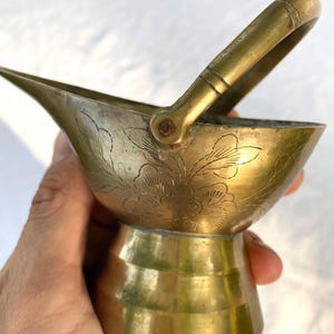 May include: A small brass coal scuttle with a polished finish, featuring an etched floral design on the body, a wide pouring spout, a sturdy pedestal base, and a hinged handle that pivots at the sides to allow for easy carrying and tilting.