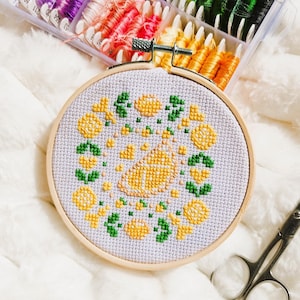 May include: A completed cross-stitch embroidery hoop featuring a yellow and green floral design. The embroidery is set in a wooden hoop, with a pair of small scissors resting nearby. Colorful embroidery floss is visible in the background.