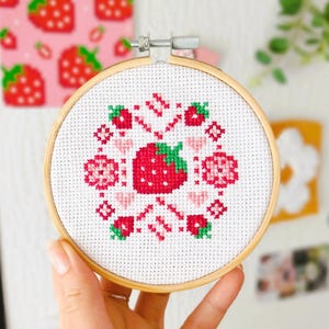 May include: A cross-stitch embroidery hoop featuring a central red strawberry with green leaves, surrounded by pink hearts and flowers. The design is set against a white fabric, framed by a wooden hoop. A pink strawberry pattern is visible in the background.
