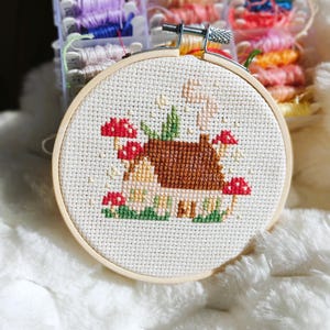 May include: A cross-stitch embroidery hoop with a mushroom house design. The house has a brown roof and is surrounded by red mushrooms and green grass. The embroidery is set in a wooden hoop, with colourful embroidery floss in the background.