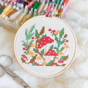 Peut inclure: Broderie au point de croix terminée dans un cercle en bois. Le motif représente des champignons rouges à pois blancs, des feuilles vertes et des baies. Des ciseaux argentés sont posés près du cercle. Un récipient en plastique transparent rempli de fil à broder coloré est en arrière-plan.