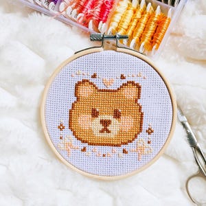 Peut inclure: Un cercle de broderie au point de croix avec un motif d'ours brun sur fond bleu clair. L'ours a un visage brun clair et un nez brun foncé. Des fils à broder de différentes couleurs et des ciseaux sont également visibles.
