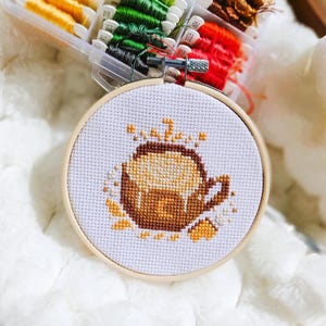 Peut inclure: Un cercle de broderie au point de croix avec un motif de tasse à café marron et beige. La broderie est placée dans un cercle en bois, avec un fond en tissu blanc. Un récipient en plastique transparent de fil à broder coloré est en arrière-plan.