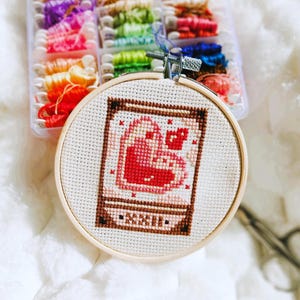 Peut inclure: Un projet de point de croix terminé dans un cercle à broder en bois, représentant un cœur rouge et une tasse à café. Un récipient en plastique transparent rempli de fil à broder coloré est en arrière-plan.