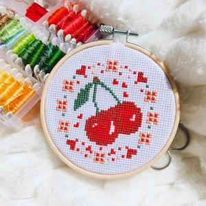 Peut inclure: Un cercle de broderie au point de croix avec un motif de deux cerises rouges et des feuilles vertes. Le motif est entouré de petits motifs floraux rouges et orange. La broderie est placée dans un cercle en bois, avec des bobines de fil à broder coloré en arrière-plan.