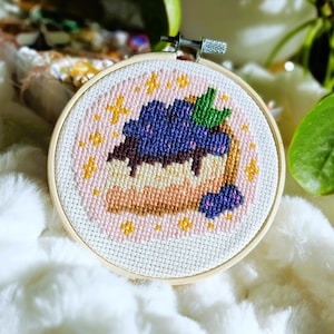 Peut inclure: Un cercle de broderie au point de croix représentant une part de cheesecake aux myrtilles. Le motif comprend un fond rose clair avec des étoiles jaunes. La part de cheesecake a une croûte brune, une garniture blanche et des myrtilles bleues et du chocolat.