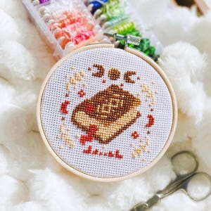 Peut inclure: Un cercle de broderie au point de croix avec un motif de livre marron et des accents rouges, avec le mot "books" brodé en rouge. La broderie est réalisée sur un tissu blanc, dans un cercle en bois. Des fils à broder et des ciseaux sont également visibles.