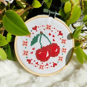 Peut inclure: Un cercle de broderie au point de croix représentant une paire de cerises rouges avec des feuilles vertes. Le motif est entouré d'accents floraux rouges et roses sur fond blanc. Le cercle est dans un cadre en bois.