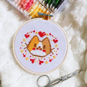 Peut inclure: Un cercle de broderie au point de croix avec un motif de chien corgi, entouré de cœurs rouges et d'empreintes de pattes jaunes. Le corgi a un pelage brun et beige avec un museau blanc. Fils à broder et ciseaux en arrière-plan.