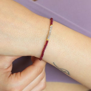May include: A delicate beaded bracelet with small, faceted red beads, grey beads and gold-coloured accents. The bracelet is worn on a wrist.