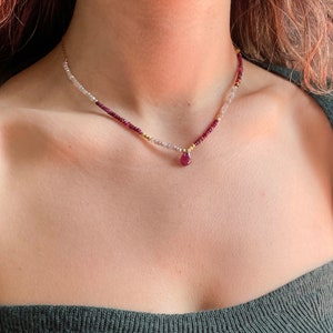 Op de afbeelding: Een delicate ketting met kleine, lichtgekleurde kralen, afgewisseld met robijnkleurige kralen en gouden accenten. Een druppelvormige robijn hanger hangt in het midden. De ketting wordt gedragen over een donkergroene top.