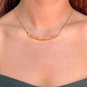 Può includere: Una delicata collana con una fila di piccole pietre preziose di opale sfaccettate nei toni del giallo e dell'arancione. Gli opali sono infilati su una sottile catena dorata. La collana è indossata intorno al collo.