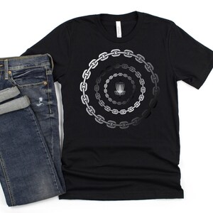 K&ouml;nnte beinhalten: Schwarzes T-Shirt mit einem Motiv eines Disc-Golf-Korbs, der von Ketten umgeben ist. Die Ketten sind kreisf&ouml;rmig angeordnet und verlaufen von dunkelgrau zu hellgrau.