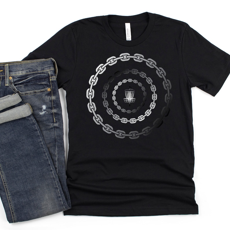 K&ouml;nnte beinhalten: Schwarzes T-Shirt mit einem Motiv eines Disc-Golf-Korbs, der von Ketten umgeben ist. Die Ketten sind kreisf&ouml;rmig angeordnet und verlaufen von dunkelgrau zu hellgrau.