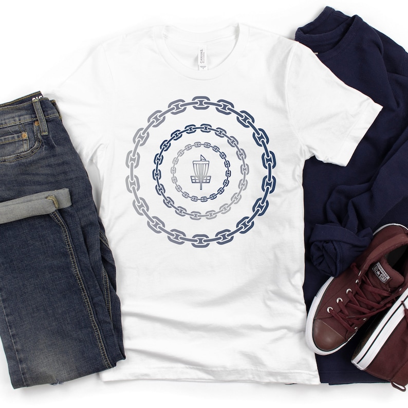 K&ouml;nnte beinhalten: Wei&szlig;es T-Shirt mit einer blauen und grauen Grafik eines Disc-Golf-Korbs, der von Ketten umgeben ist. Das Shirt liegt auf einer wei&szlig;en Oberfl&auml;che mit einer blauen Jeans, einem marineblauen Pullover und braunen Turnschuhen.