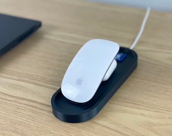 Puede incluir: Un ratón inalámbrico blanco con un logotipo de Apple se encuentra sobre una base de carga negra. La base está sobre una superficie de madera.