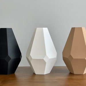 Puede incluir: Tres jarrones geométricos en negro, blanco y beige. Los jarrones tienen todos la misma forma, pero diferentes colores. Están hechos de un material liso y mate.