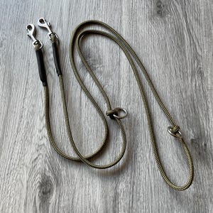 May include: An olive green braided rope lead featuring two silver-toned metal swivel snap hooks at one end, secured with black sleeves, and two silver-toned metal rings integrated along the length of the rope for adjustable length configurations.