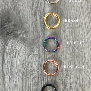 May include: Five circular metal rings arranged vertically. The top ring is silver-colored labeled STAINLESS STEEL, followed by a gold-colored ring labeled BRASS, a multi-colored iridescent ring labeled JET FUEL, a copper-colored ring labeled ROSE GOLD, and a black ring labeled BLACK.