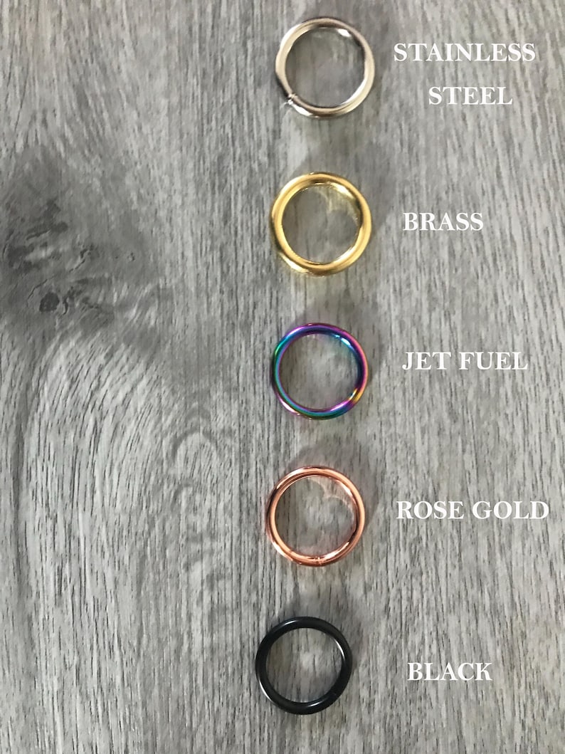May include: Five circular metal rings arranged vertically. The top ring is silver-colored labeled STAINLESS STEEL, followed by a gold-colored ring labeled BRASS, a multi-colored iridescent ring labeled JET FUEL, a copper-colored ring labeled ROSE GOLD, and a black ring labeled BLACK.