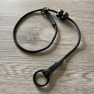 May include: A black cord lanyard featuring a circular metal ring at one end, secured with a black heat-shrink sleeve. The cord includes two adjustable black plastic cord locks and a small metal loop, creating a continuous loop design for utility.