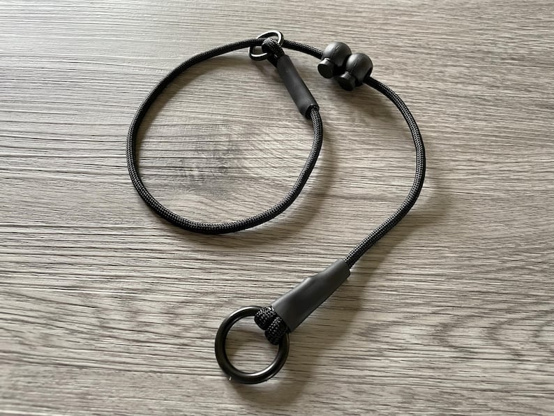 May include: A black cord lanyard featuring a circular metal ring at one end, secured with a black heat-shrink sleeve. The cord includes two adjustable black plastic cord locks and a small metal loop, creating a continuous loop design for utility.