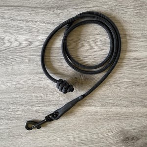 May include: Black dog lead made from a robust, woven material. It has a black metal clasp and a knotted handle for a secure grip. The lead is coiled on a wooden surface.