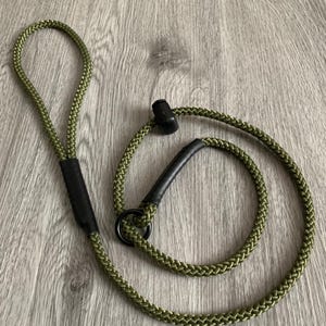 May include: An olive green braided rope leash featuring a black circular metal ring, a black sliding stopper, and black reinforced sleeves at the handle and attachment points. The durable cord construction provides a flexible design.