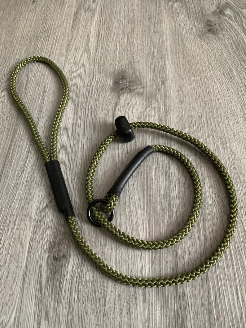 May include: An olive green braided rope leash featuring a black circular metal ring, a black sliding stopper, and black reinforced sleeves at the handle and attachment points. The durable cord construction provides a flexible design.