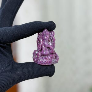 Könnte beinhalten: Eine kleine, aufwendig geschnitzte Figur der hinduistischen Gottheit Ganesha. Die Statue hat eine tiefe, satte lila Farbe und scheint aus Stein zu sein. Die Figur wird in einer schwarz behandschuhten Hand gehalten, mit unscharfem Hintergrund.