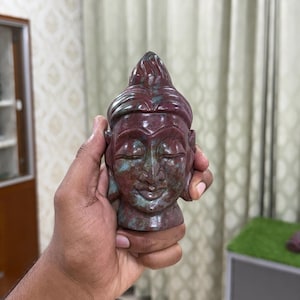 以下が含まれることがあります： 赤と緑の色合いの、彫刻された仏陀の頭の彫刻。彫刻は目を閉じ、穏やかな表情をしています。頭は手で持たれており、石の彫刻の複雑な詳細を示しています。