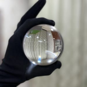 May include: A clear glass sphere held in a black gloved hand. The sphere reflects a room with patterned curtains, a brown door, and a window. The sphere is transparent and captures the surrounding environment.