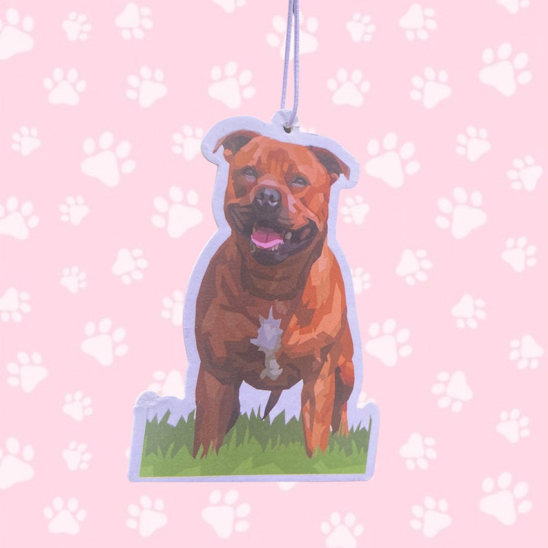 May include: A brown dog air freshener with a smiling face, hanging from a white string. The dog is depicted in a low-poly style, with green grass at the bottom. The background is pink with white paw prints.