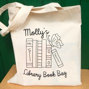 May include: A white canvas tote bag with black text and a graphic of a stack of books and a butterfly. The text reads "Molly's Library Book Bag".