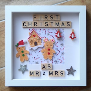 Puede incluir: Un marco de fotos blanco con una casa de jengibre, figuras de jengibre y un árbol de Navidad. El marco tiene las palabras "FIRST CHRISTMAS AS MR & MRS" deletreadas con fichas de letras.
