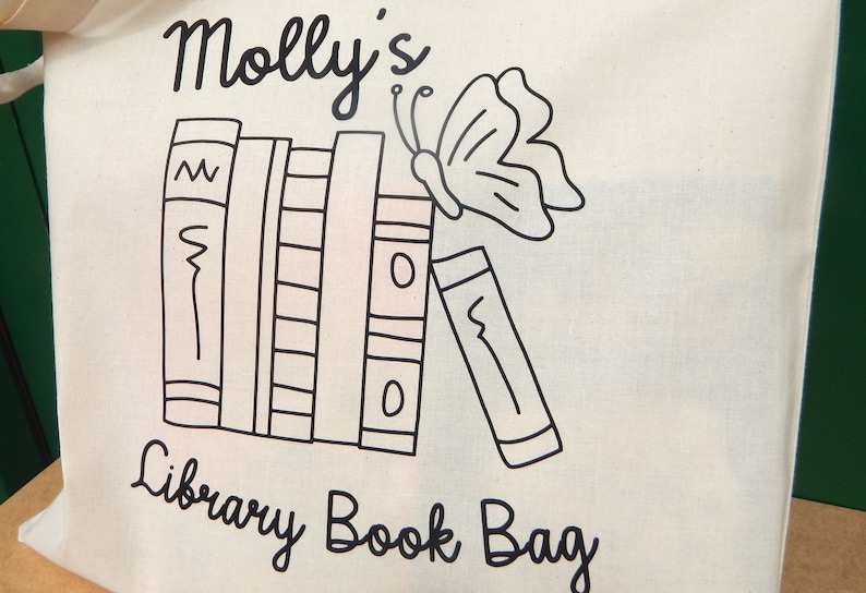 May include: A white canvas tote bag with black text and a black and white illustration of a stack of books and a butterfly. The text on the bag reads "Molly's Library Book Bag".