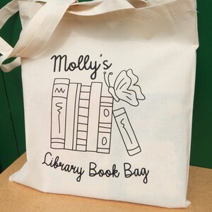 May include: White canvas tote bag with black text and a graphic of a stack of books and a butterfly. The text reads "Molly's Library Book Bag".