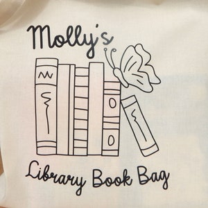 May include: A white canvas tote bag with black text and a black and white illustration of a stack of books and a butterfly. The text reads "Molly's Library Book Bag".