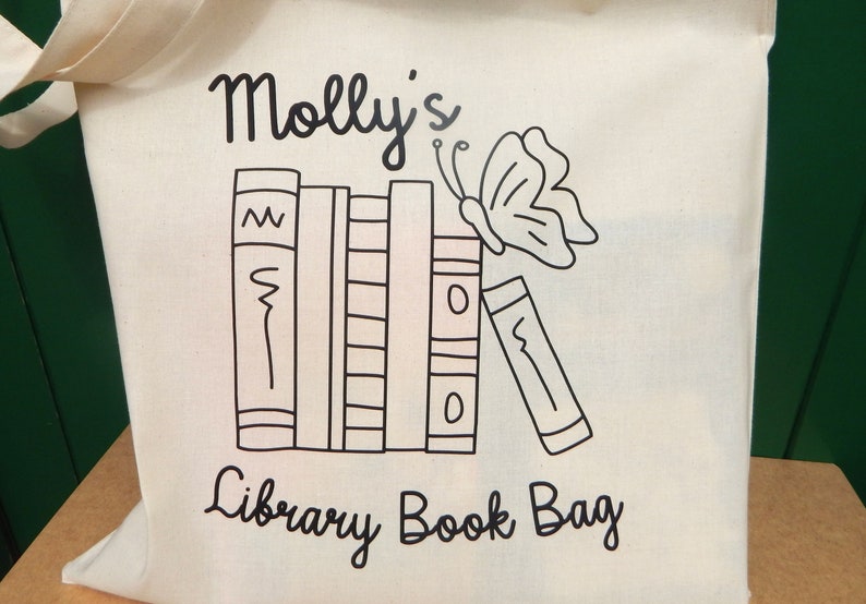 May include: A white canvas tote bag with black text and a black and white illustration of a stack of books and a butterfly. The text reads "Molly's Library Book Bag".